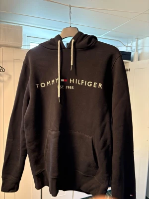 Svart hoodie från Tommy Hilfiger - Svart hoodie från Tommy Hilfiger med vit logotyp och text på bröstet samt klassiska Hilfiger-färger. Huva med vita snören och stor magficka framtill. Mjuk bomullskänsla och ribbade muddar vid ärmslut och nederkant. Perfekt för en avslappnad look.
