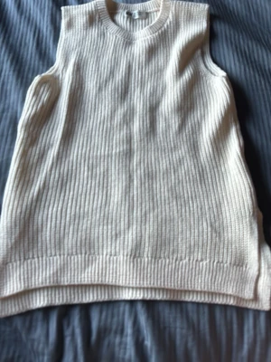 Beige ribbad stickad väst - Säljer en beige stickad väst med ribbad struktur och rund hals. Perfekt att styla över en skjorta eller t-shirt för en trendig lager-på-lager-look. Västen har en loose passform och är ärmlös.