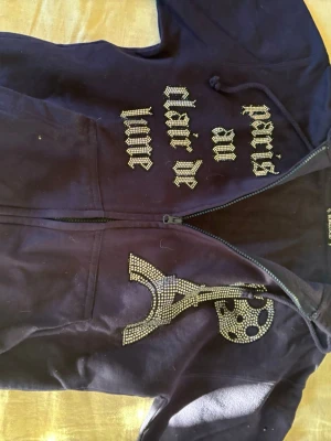 Lila hoodie med strass och tryck - Säljer en lila hoodie från Bik Bok med dragkedja och huva. Framsidan har texten 'Paris au clair de lune' i strass och en stor Eiffeltorn-design i strass på ena sidan. Hoodien har fickor framtill och är gjord i mjukt bomullsmaterial. Perfekt för dig som gillar statement-plagg.