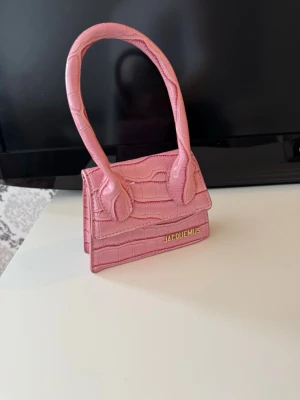 Jacquemus Le Chiquito handväska rosa, axelrem ingår! - Axelrem ingår! Den ikoniska Jacquemus Le Chiquito handväska i ljusrosa med krokopräglat mönster. Väskan har ett topphandtag och guldfärgad logga framtill. Perfekt för dig som gillar trendiga accessoarer och vill sticka ut med en unik miniväska.
