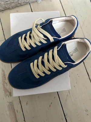 Blå Maison Margiela sneakers - Snygga blå sneakers från Maison Margiela med beige snören och gummisula. Ovandelen är i en mix av mocka och slätt läder, vilket ger en schysst kontrast. Insidan är vit och sulan har en klassisk brun färg. Perfekta för dig som gillar stilrena men unika skor. Lite små i storlek så passar 43/42