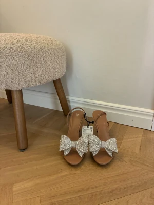 Bruna klackar med strassrosetter - Säljer ett par bruna pumps med öppen tå och transparent rem över foten. Skorna har en hög smal klack och är dekorerade med stora glittriga rosetter i strass framtill. Märket är Zara, prislapp kvar. Nypris 560 kr.