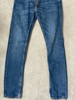 Blå jeans från Nudie Jeans - Säljer ett par klassiska blå jeans från Nudie Jeans med raka ben och snygga kontrastsömmar. Jeansen har fem fickor och den ikoniska broderade vågen på bakfickan. Tillverkade i slitstark denim med en cool tvättad look. Waist 29, length 32