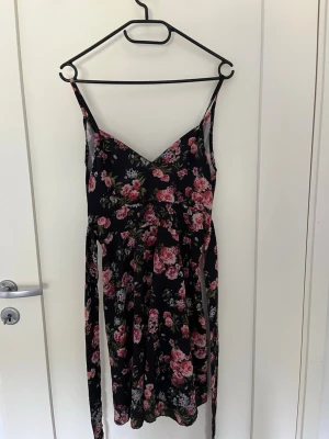 Svart blommig sommarklänning - Säljer en svart klänning med tunna axelband och v-ringning. Klänningen har ett blommigt mönster i rosa och grönt, samt en lätt utställd kjol. Perfekt för varma dagar och har en feminin, flowy siluett.