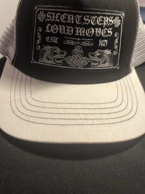 Silent Steps Loud Moves truckerkeps - Snygg svart och vit truckerkeps med mesh på sidorna och broderad patch framtill med texten 'Silent Steps Loud Moves' och coola grafiska detaljer. Justerbar baktill för perfekt passform. Perfekt för dig som vill sticka ut med en unik keps.