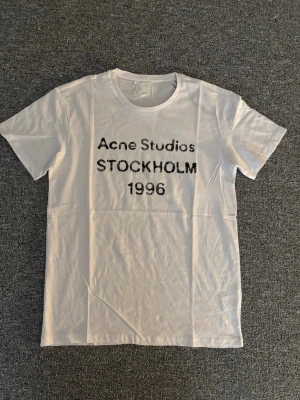 Vit Acne Studios t-shirt - Säljer en vit t-shirt från Acne Studios med svart text 'Acne Studios STOCKHOLM 1996' på bröstet. Klassisk rund halsringning och korta ärmar. Tillverkad i mjuk bomull för en clean och stilren look. ALLA LAPPAR SAKNAS DÄRAV PRISET