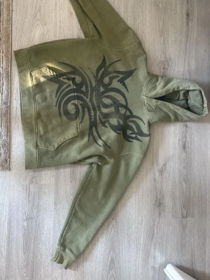 Olivgrön hoodie med tribaltryck - Säljer en olivgrön hoodie med stort svart tribaltryck och små silvriga nitar på framsidan. Hoodien har en klassisk känguruficka och huva. Perfekt för dig som gillar streetwear och vill sticka ut lite extra. Materialet känns mjukt och bekvämt.