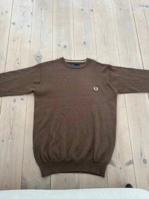 Brun stickad tröja från Fred Perry - Snygg brun stickad tröja från Fred Perry med rund halsringning och diskret logga broderad på bröstet. Tröjan har långa ärmar och ribbade muddar vid ärmslut och nederkant. Perfekt för lager på lager och enkel att matcha.