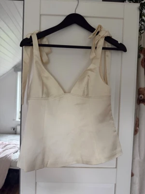 Ljusbeige linnetopp från Gina Tricot - Supersöt ljusbeige linnetopp från Gina Tricot med knytbara axelband och lätt v-ringad hals. Tillverkad i ett glansigt satintyg som ger en lyxig känsla. Perfekt att styla med jeans eller kjol för en trendig look.