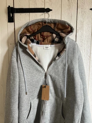 Grå zip hoodie från Burberry - Snygg grå zip hoodie från Burberry med klassiskt rutigt foder i huvan och vita dragsnören. Hoodien har fickor framtill och är tillverkad i mjuk bomullsblandning. Perfekt för dig som vill ha en stilren och exklusiv look med en twist av Burberrys ikoniska mönster. Den är i storlek L men passar som en M/L. Den har mönster inne och i luvan. Skicka vid frågor och pris går att diskutera👍