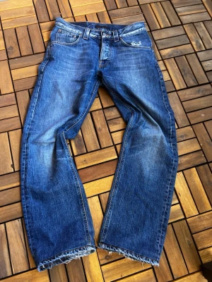 Blå raka jeans med rå kant - Säljer ett par blå jeans med rak passform med en liten flare på slutet , fransig kant nedtill. Jeansen har klassisk femficksdesign, slitningar och snygg tvätt för en avslappnad look. Perfekta för dig som gillar en lite mer edgy stil.