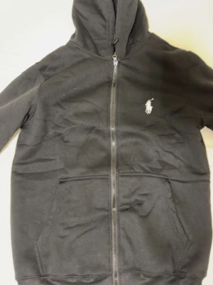 Svart hoodie från Polo Ralph Lauren - Säljer en svart hoodie med dragkedja från Polo Ralph Lauren. Klassisk design med vit broderad logga på bröstet, kängurufickor och ribbade muddar. Tillverkad i mjukt bomullsmaterial och har en skön huva med dragsko.