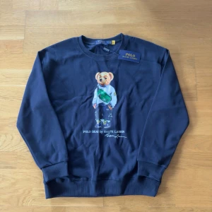 Mörkblå sweatshirt Polo Ralph Lauren - Mörkblå sweatshirt från Polo Ralph Lauren med ikoniska Polo Bear-trycket framtill. Rund halsringning och ribbade muddar vid ärmslut och nederkant. Perfekt för en avslappnad och trendig stil.