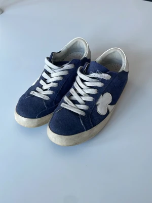 Blå sneakers från The Gallerist - Denna skon tillverkas inte så den är limited edition och det finns inte långa i samma storlek. Snygga blå sneakers från The Galleryist med vit sula och vita detaljer. Skorna har en unik klöverdetalj på sidan och är gjorda i mocka material. Klassisk låg modell med en RIKTIGT skön innersula som det känns som att man går på moln.