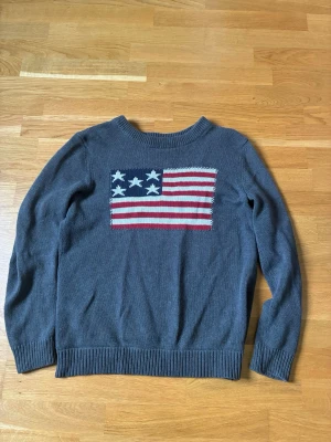 Grå stickad tröja med flaggmotiv - Stickad tröja i mörkgrått från Selected Homme med ett stort amerikanskt flaggmotiv på bröstet. Tröjan har rund halsringning och ribbade muddar vid ärmslut och nederkant. Perfekt för dig som gillar en casual och cool stil.