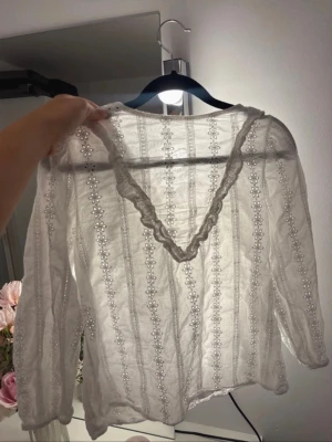Vit broderad blus med spetsdetaljer - Söt vit blus med broderade blommönster och spetskantad V-ringning. Blusen har långa ärmar och är gjord i ett tunt, lätt material som ger en romantisk och luftig känsla. Perfekt för dig som gillar en feminin och trendig stil.