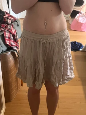 Beige volangkjol med spetskant - Söt beige kortkjol med volanger och spetskant längst ner. Kjolen har resår i midjan och en liten knytning framtill. Perfekt för varma dagar och ger en avslappnad men ändå gullig look. Knappt använd och inget slitage. 