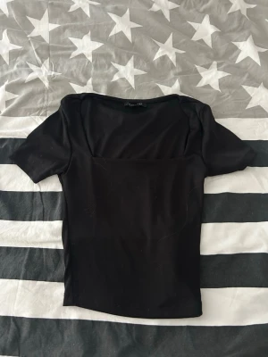 Svart topp med fyrkantig ringning - Stilren svart topp med fyrkantig ringning och korta ärmar. Toppen har en slim passform och är tillverkad i ett mjukt, stretchigt material som känns bekvämt mot huden. Perfekt att matcha med både jeans och kjol.