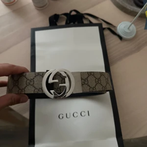 Mönstrat bälte från Gucci - Säljer ett stilrent bälte från Gucci med det klassiska GG-mönstret. Bältet har en stor, rund spänne i silverfärg med Gucci-logotypen. Perfekt för att ge en lyxig touch till din outfit. Garanterat äkta. Tveka inte på att höra av dig ifall du har frågor! Obs endast produkten ingår 