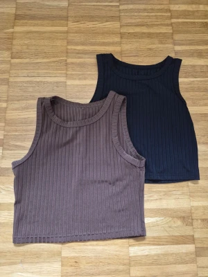 Ribbad brun och svart linnetopp - Två ribbade linnetoppar i brunt och svart. Båda är ärmlösa och har en croppad passform med rund halsringning. Perfekta att matcha med jeans eller kjol för en enkel och clean look. Säljer både dessa tsm!