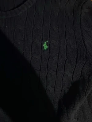Svart kabelstickad tröja Polo Ralph Lauren - Kabelstickad svart tröja från Polo Ralph Lauren med klassisk grön logga broderad på bröstet. Tröjan har rund halsringning och ribbade muddar nedtill. Perfekt för dig som gillar stilrena och tidlösa plagg.