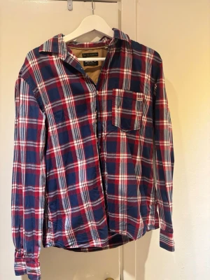 Rutig skjorta från Jack & Jones - Säljer en klassisk rutig skjorta från Jack & Jones Vintage Clothing i blått, rött och vitt. Skjortan är långärmad, har en bröstficka och är tillverkad i mjuk bomull. Perfekt för en avslappnad och stilren look.