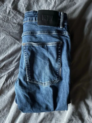 Blå slim jeans från Neuw - Säljer ett par blå skinny jeans från Neuw med klassisk femficksdesign och svart patch bak i midjan. Jeansen har smal passform och är tillverkade i mjukt denimtyg med snygga kontrastsömmar. Perfekta för en trendig och avslappnad stil.