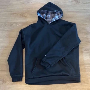 Svart hoodie med rutigt foder - Säljer en svart hoodie med rutigt innerfoder i huvan och nederkanten. Hoodien har en klassisk passform, långa ärmar och en liten vit brodyrdetalj på ärmen. Perfekt för dig som gillar en enkel men ändå unik stil.