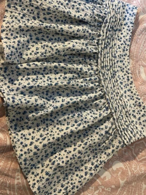 Vit minikjol med blå blommor - Supersöt minikjol i vitt med blått blommönster och bred smock i midjan. Kjolen har en luftig och volangig form som ger en avslappnad och trendig look. Perfekt för sommardagar och enkel att matcha med en enkel topp. Materialet känns mjukt och lätt.