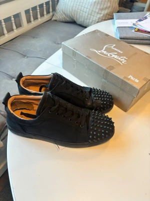 Svarta Louboutin sneakers med nitar - Säljer ett par svarta sneakers från Christian Louboutin med coola svarta nitar över tån. Skorna har lågt skaft, svarta skosnören och en klassisk röd insida. Kvitto och äkthetsbevis finns. Mycket slitet skick. Det finns extra nitar i förpackningen att sätta på. En skomakare kan laga sprickorna. Säljes mycket billigt pga detta! 