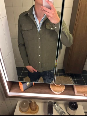 Olivgrön overshirt med knappar - Säljer en olivgrön overshirt i 100% polyester med klassisk krage och vita knappar framtill. Skjortan har två bröstfickor med lock och knapp, perfekt för en avslappnad men snygg stil. Passar till lager-på-lager och funkar året runt.