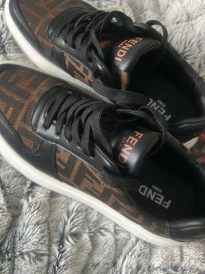 Fendi bruna och svarta sneakers - Snygga sneakers med klassiskt FF-mönster i brunt och svart. Skorna har svarta läderdetaljer, vita sulor och Fendi-logga på plös och häl. Tjocka svarta skosnören och en modern, chunky design. Perfekta för dig som vill sticka ut med lyxig streetstyle. Helt nya inte använda