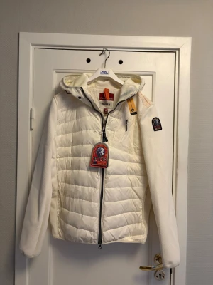 Parajumper cardigan - Säljer en snygg vit pufferjacka från Parajumpers med huva och dragkedja framtill. Jackan har quiltad front och rygg, samt släta ärmar. Märkets logga sitter på vänster ärm och jackan har en gul detalj vid halsen. Perfekt för vinterdagar, vårdagar samt höstdagar