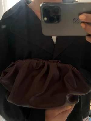Vinröd clutchväska med axelrem - Säljer en vinröd clutchväska i mjukt, slätt material med rynkad design och lång axelrem. Väskan har en modern, puffig form och kan bäras både i handen och över axeln. Perfekt för dig som gillar trendiga accessoarer med unik look.