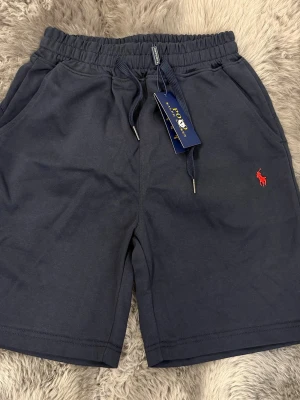 Mörkblå shorts från Polo Ralph Lauren - Snygga mörkblå shorts från Polo Ralph Lauren med klassisk röd broderad logga på benet. Shortsen har dragsko i midjan och två sidofickor. Perfekta för sommardagar och chill häng.