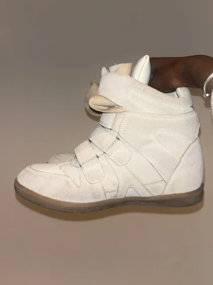 Gina Isabel marant sneakers - Snygga vita sneakers i mocka med högt skaft och flera breda kardborreband. Skorna har en chunky sula i gummi och vadderad plös för extra komfort. Perfekta för dig som gillar streetstyle och vill sticka ut med en unik look.