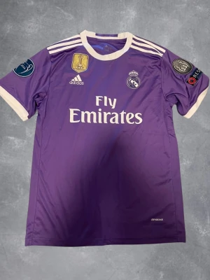 Lila Real Madrid Ronaldo matchtröja - Snygg lila Real Madrid matchtröja från Adidas med vita detaljer och tryck. Tröjan har klubbmärke, sponsorlogga och Champions League-patchar på ärmarna. På ryggen står det Ronaldo och nummer 7. Tillverkad i lätt funktionsmaterial.