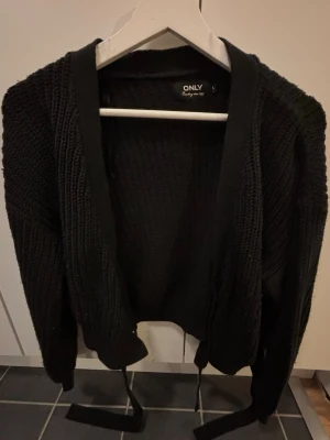 Svart stickad kofta från ONLY - Snygg svart stickad kofta från ONLY med öppen front och breda ribbade kanter. Koftan har långa ärmar och ett bälte i midjan för en avslappnad look. Perfekt att slänga över en t-shirt eller topp för extra värme.