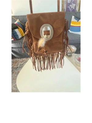 Brun mocka axelremsväska med fransar - Säljer en brun handväska i mocka med långa fransar och axelrem. Väskan har ett dekorativt spänne framtill med en fjäderdetalj. Perfekt för dig som gillar boho-stil och vill ha något unikt till din outfit.