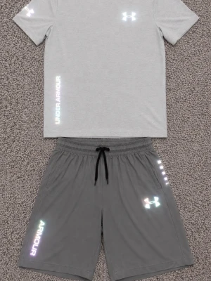 Grått träningsset från Under Armour - Snyggt träningsset från Under Armour med en ljusgrå t-shirt och matchande mörkgrå shorts. T-shirten har reflekterande logga och textdetaljer, medan shortsen har dragsko i midjan och reflekterande logga på benet. Perfekt för gymmet eller löprundan.