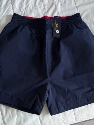 Ralph Lauren Badbyxor - Snygga marinblå badbyxor från Polo Ralph Lauren med elastisk midja och röd insida. Klassisk modell med sidofickor och en bakficka. Tillverkade i snabbtorkande material, perfekta för stranden eller poolen. Helt nya. Säljer då de inte passar.