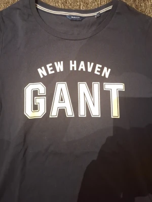 Mörkblå långärmad GANT t-shirt - Snygg mörkblå långärmad t-shirt från GANT med trycket 'NEW HAVEN GANT' i vitt framtill. Rund halsringning och klassisk passform. Perfekt för en avslappnad och sportig stil.
