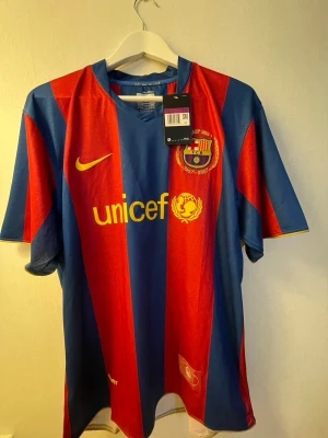 Barcelona Ronaldinho matchtröja Nike - Säljer en klassisk FC Barcelona matchtröja från Nike med Ronaldinho och nummer 10 på ryggen. Tröjan har breda blå och röda ränder, gul Nike-logga och Unicef-tryck på bröstet. Tillverkad i lätt och ventilerande polyester, perfekt för fotboll eller samling.