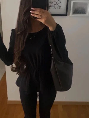 Svart långärmad blus med markerad midja - Snygg svart blus med lång ärm och markerad midja som ger en feminin siluett. Blusen har rund halsringning och är perfekt att styla med både jeans och kjol. Enkel men stilren design som passar till många olika looks.