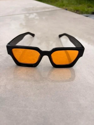 Svarta solglasögon med orange glas - Snygga solglasögon med svart, kantig båge och orangefärgade glas. Glasögonen har en modern, retroinspirerad look och passar perfekt för dig som vill sticka ut. Bågarna är i plast och glasen ger cool kontrast till din outfit.🕶️☀️⛱️