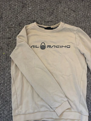 Beige Sail Racing sweatshirt - Beige sweatshirt från Sail Racing med svart logotyptryck över bröstet. Tröjan har rund halsringning och långa ärmar. Perfekt för en avslappnad och stilren look.