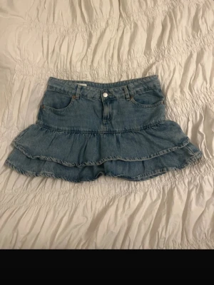 Blå volangkjol i denim från Zara - Säljer en blå jeanskjol från Zara med dubbla volanger och klassisk femficksdesign. Kjolen har en kort modell och är gjord i bomull. Perfekt för dig som gillar en avslappnad men trendig stil.