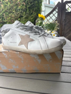 Vita sneakers från Golden Goose - Säljer ett par vita Golden Goose sneakers med grå snörning och beige stjärndetalj på sidan. Skorna har låg profil och är tillverkade i skinn med en klassisk rund tå och platt sula. Perfekta för dig som gillar en clean men ändå unik look.