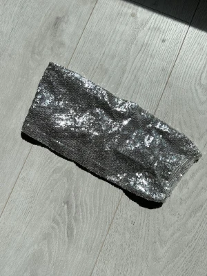 Silvrig paljett bandeau tubtopp - Glittrig bandeau tubtopp i silver med paljetter över hela tyget. Perfekt för dig som vill sticka ut och glänsa. Toppen är axelbandslös och har en tajt passform som framhäver din stil. Perfekt för fest, festival, sommar!
