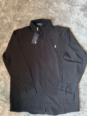Svart skjorta från Polo Ralph Lauren - Säljer en svart skjorta från Polo Ralph Lauren med klassisk krage och knappar framtill. Skjortan har lång ärm och ikonisk broderad logga på bröstet. Perfekt för dig som vill ha en stilren och tidlös look.
